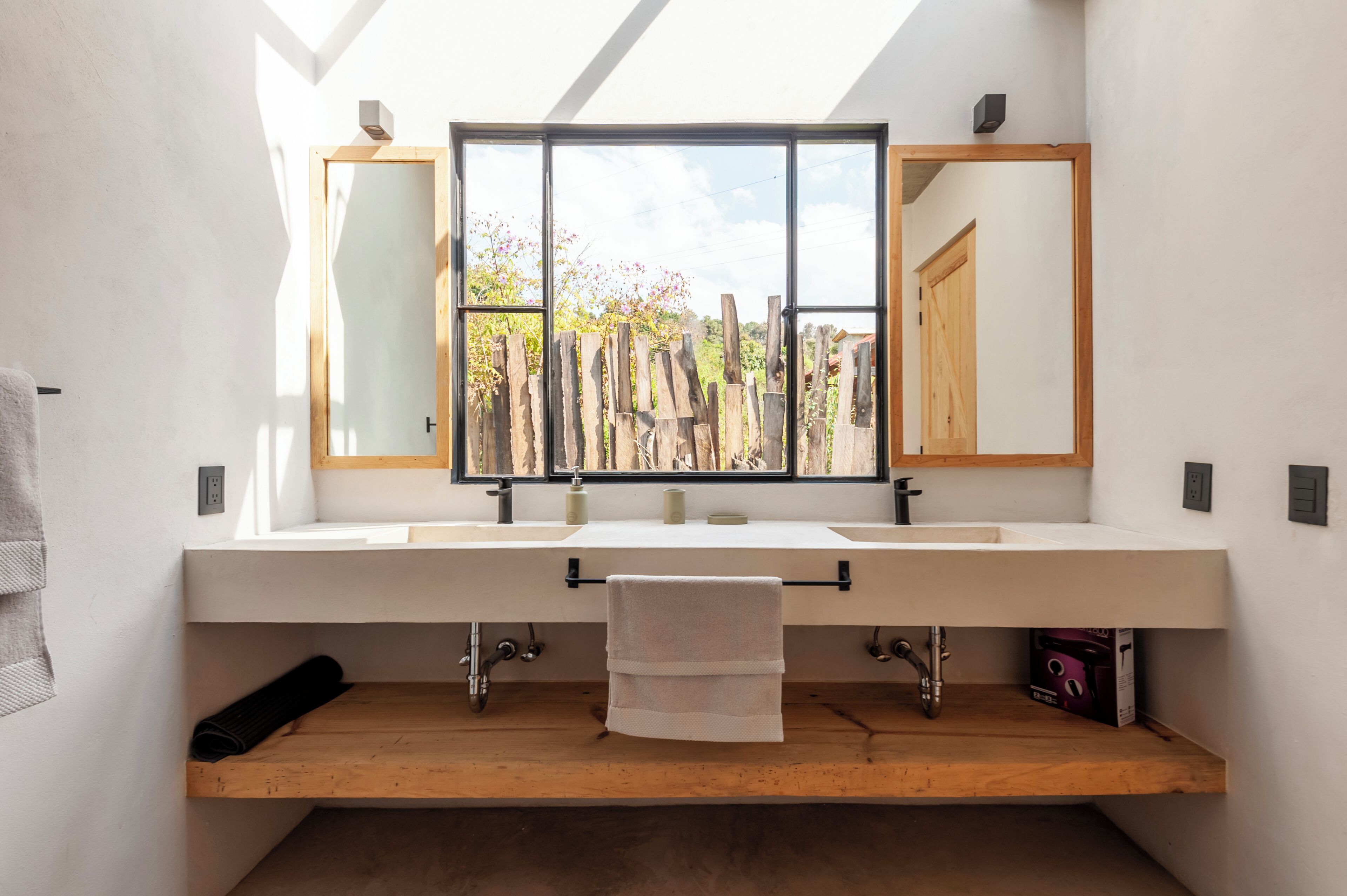A modern bathroom with a large window, wooden bench, and dual sinks.