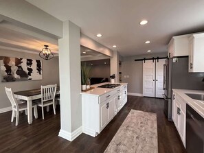 A modern kitchen with white cabinetry, a dining area with a wooden table and chairs, and a large island with a stove. - River Access•Game room•Backyard•King•W/D•Parking (Yorkville)
