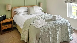 Een net opgemaakt bed met een gewatteerde spreideken, een nachtkastje met een lamp en een raam met uitzicht op groen.
