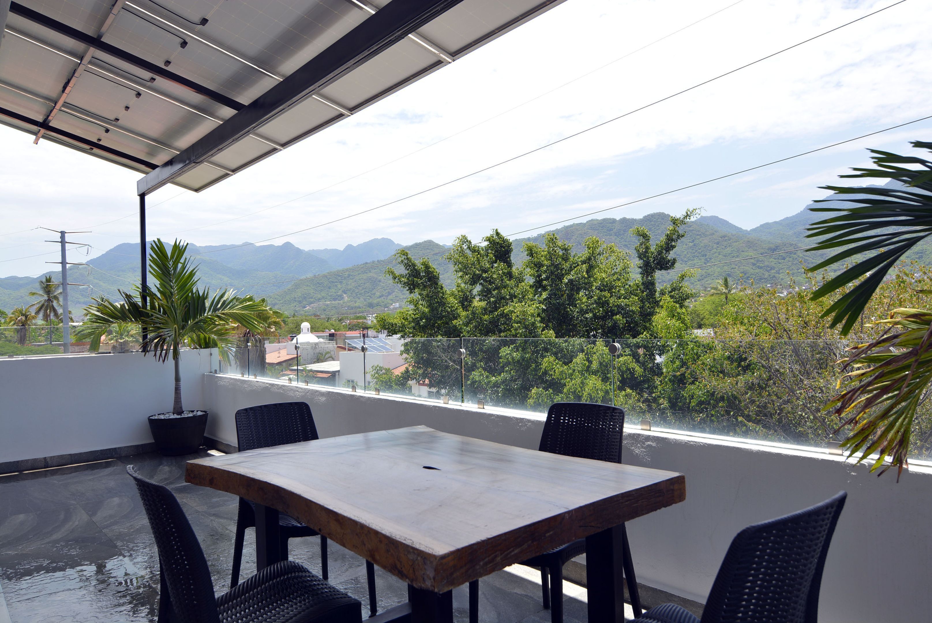 A rooftop terrace with a table and chairs, offering a view of mountains and greenery.
