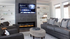Een moderne woonkamer met een open haard, een flatscreen televisie aan de muur en een grijze bank met meerdere kussens.