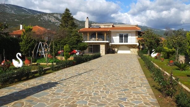 A stone pathway leading to a house with a tiled roof, surrounded by greenery and a mountainous backdrop.