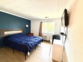 A bedroom with a bed, a desk, a television, a window, and a wall-mounted light.