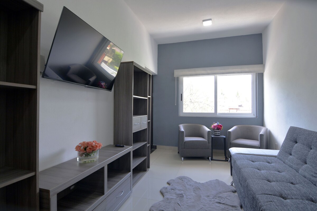 A modern living room with a flat-screen TV mounted on the wall, a grey sofa, two armchairs, a small table with a vase of flowers, and a large window providing natural light.