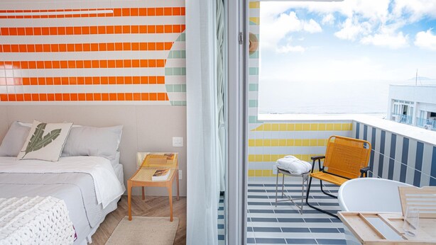 Un dormitorio moderno con cama, una mesita y una silla. Hay un balcón con vista al mar y un edificio en la distancia.