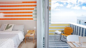 A modern bedroom with a bed, a small table, and a chair. There is a balcony with a view of the sea and a building in the distance. - Sea View and Balcony With Bathtub PI Premium (Rio de Janeiro)