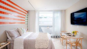 A modern hotel room with a large bed, a dining table with chairs, a TV mounted on the wall, and a view of the city through the window. - Sea View and Balcony With Bathtub PI Premium (Rio de Janeiro)