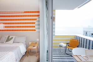 Et soveværelse med en seng, et lille bord og en stol. Der er en balkon med udsigt til havet.