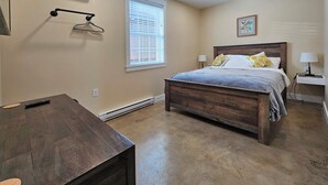 A bedroom with a wooden bed, a bedside table, a lamp, a hanging light fixture, a window with blinds, and an air conditioning unit. - Breckenridge Suites (Grove City)