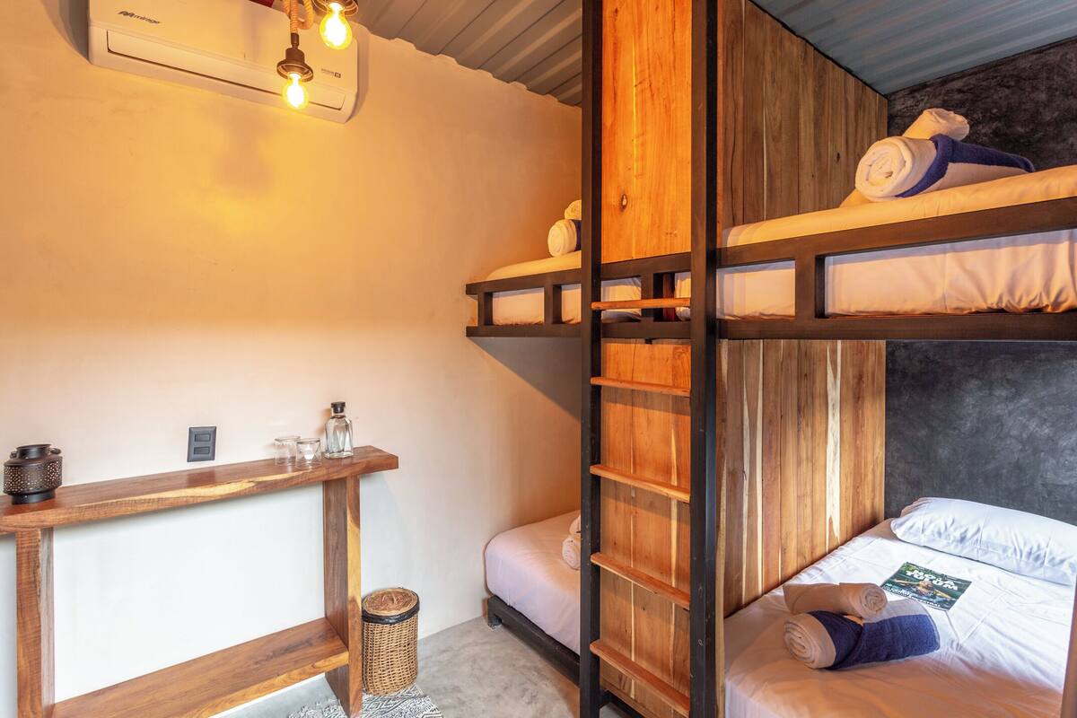 Une chambre avec un lit superposé, un bureau en bois, une petite poubelle et un climatiseur fixé au mur.
