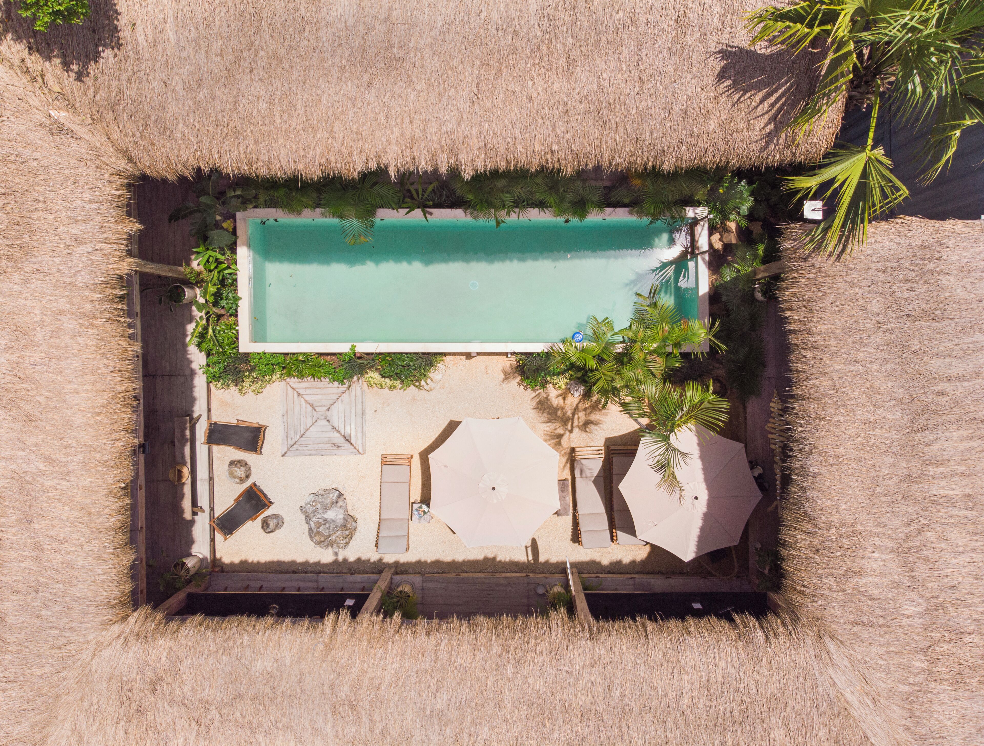 An aerial view of a square-shaped pool area with a thatched roof, surrounded by palm trees and a sandy floor.