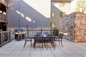 A patio with a grill, outdoor table, and chairs, a stone wall, and a building with windows.