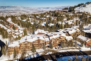 A snow-covered resort with multiple wooden buildings, likely lodges, surrounded by trees and a mountainous backdrop.