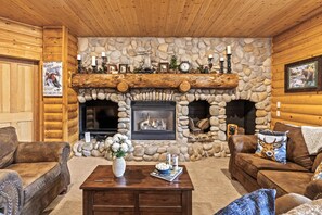 Interior de una cabaña de troncos con chimenea de piedra, muebles de madera y un reloj colgado.