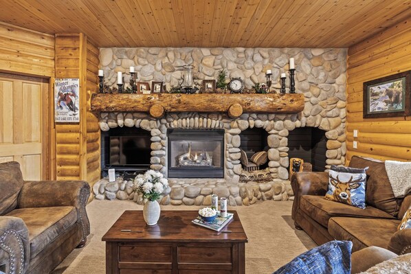 Interior de una cabaña de troncos con chimenea de piedra, muebles de cuero y una televisión.
