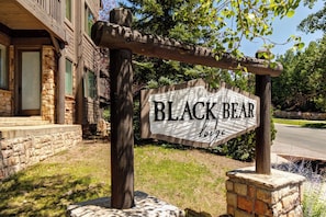 A wooden sign with 'BLACK BEAR lodge' written on it, supported by a stone base and wooden posts, with a stone building in the background.