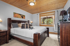 A bedroom with a wooden bed, a nightstand, a lamp, a window with curtains, and a painting on the wall.