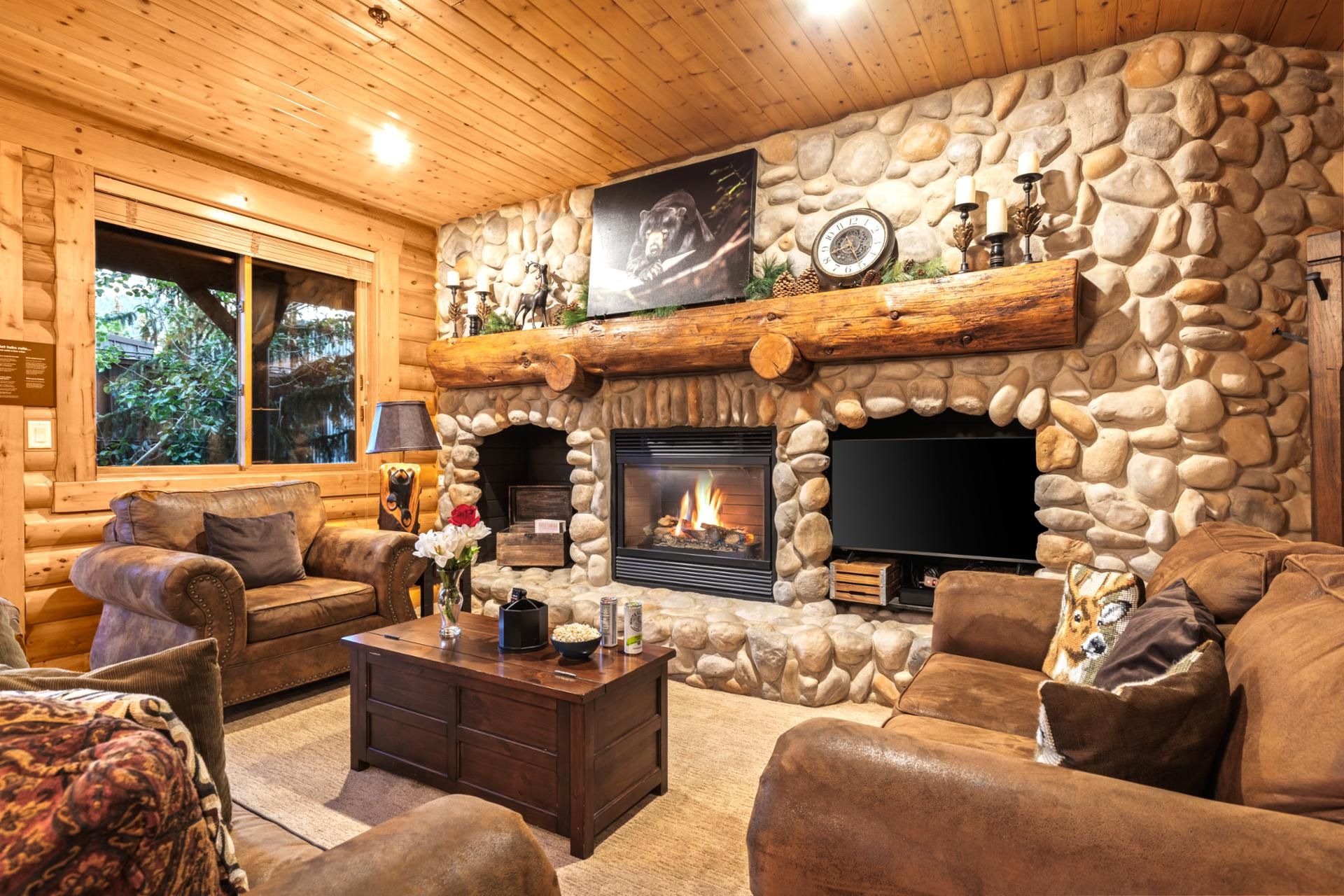 Interior de una cabaña de troncos con chimenea de piedra, muebles de cuero y un televisor sobre la chimenea.