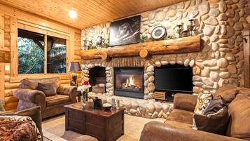 A log cabin interior with a stone fireplace, leather furniture, and a television mounted above the fireplace.