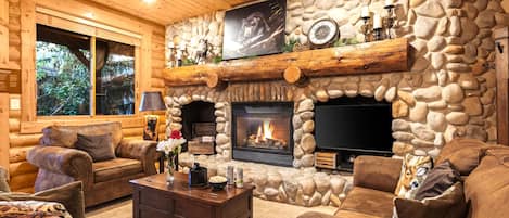 A log cabin interior with a stone fireplace, leather furniture, and a television mounted above the fireplace.