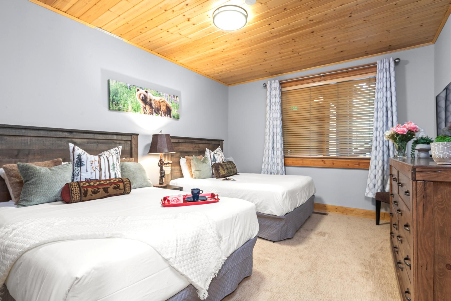 A room with two beds, a wooden ceiling, a painting of a bison, a window with blinds, and a dresser with a vase of flowers.
