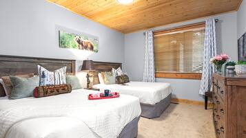 A room with two beds, a wooden ceiling, a painting of a bison, a window with blinds, and a dresser with a vase of flowers.