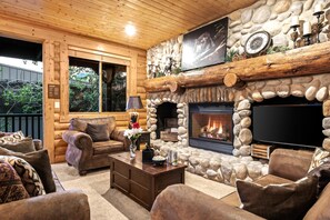 Interior de una cabaña de troncos con chimenea de piedra, muebles de madera y un televisor montado en la pared.