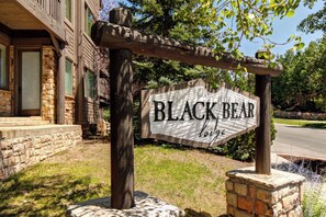 A wooden sign with 'BLACK BEAR lodge' written on it, supported by a stone base and wooden posts, with a stone building in the background.