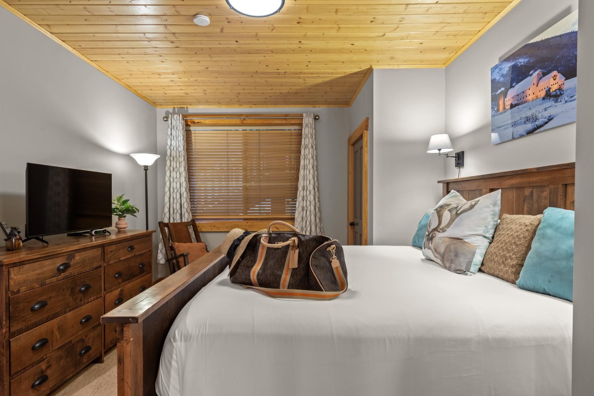 Un dormitorio con techo de madera, cama con ropa de cama blanca, televisor sobre un aparador de madera y un cuadro en la pared.