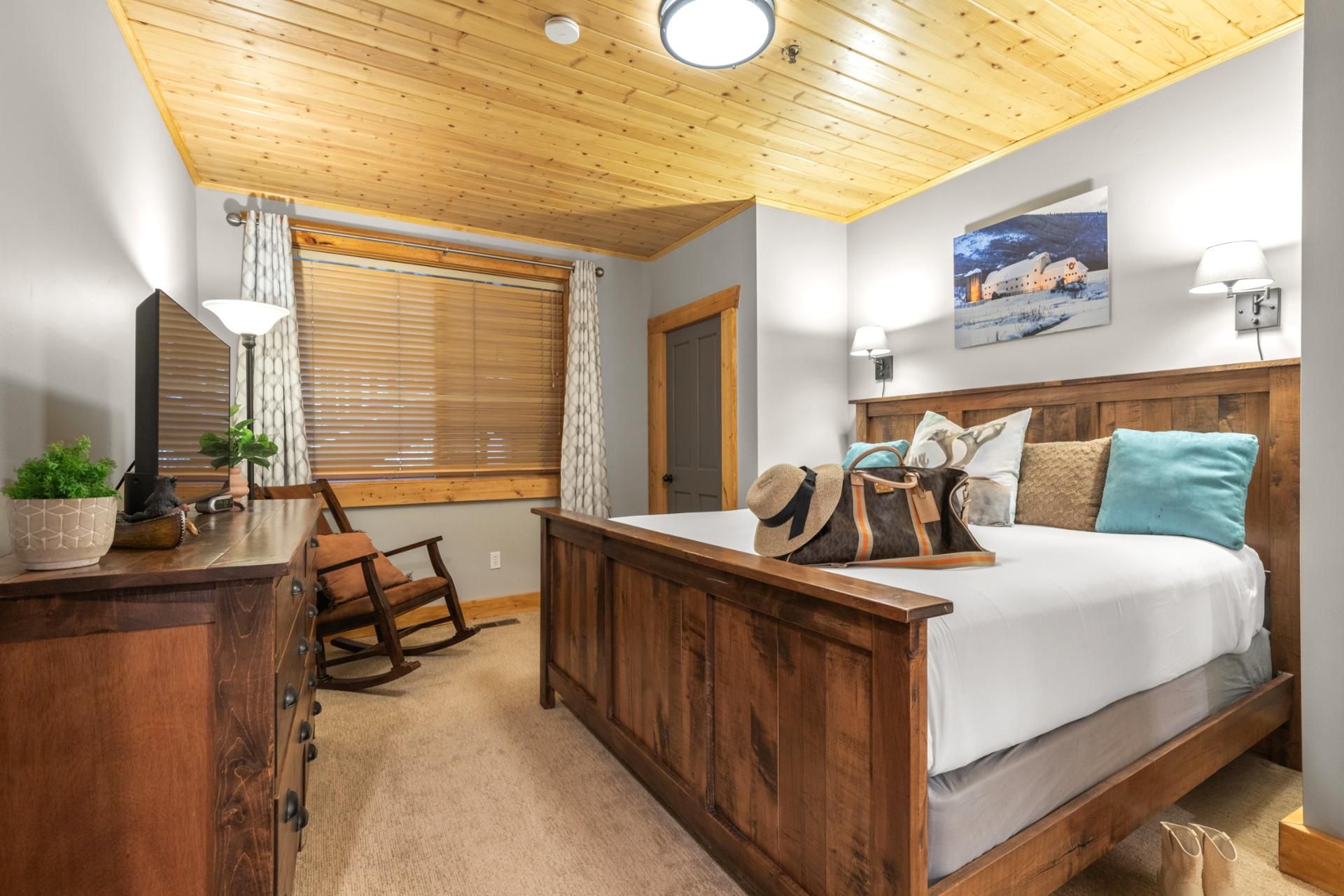 A bedroom with a wooden bed, a rocking chair, a desk with a lamp, and a window with blinds.