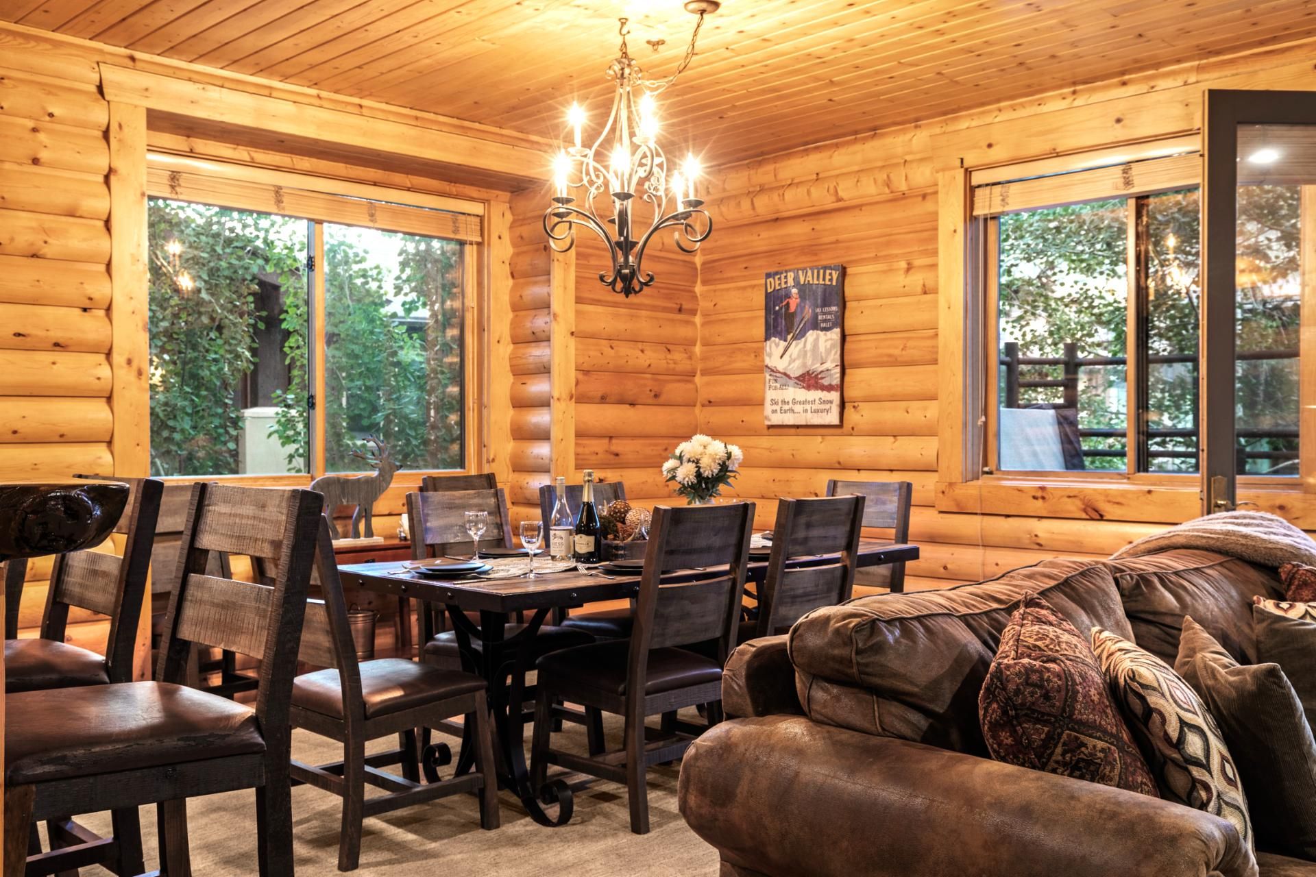 A cozy cabin interior with a dining table set for six, surrounded by wooden chairs. A chandelier hangs from the ceiling, and a poster on the wall advertises skiing.