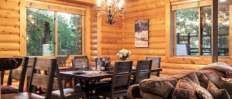 A cozy cabin interior with a dining table set for six, surrounded by wooden chairs. A chandelier hangs from the ceiling, and a poster on the wall advertises skiing.