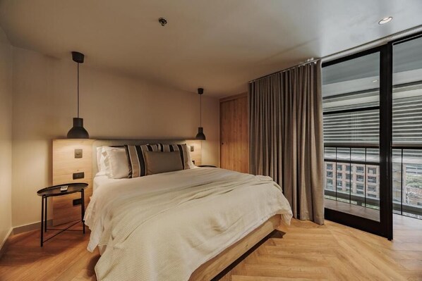 A modern bedroom with a large bed, a nightstand, a ceiling light, and a view of the city through large windows.