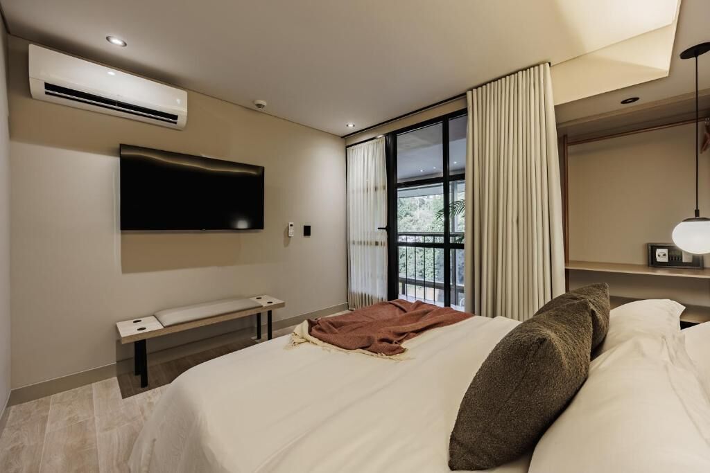 Una habitación de hotel moderna con una cama grande, un televisor de pantalla plana montado en la pared, una mesita de noche y una ventana con cortinas.