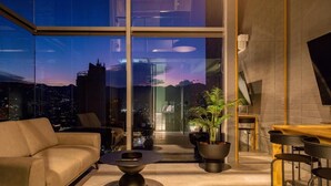 A modern living room with a sofa, a coffee table, and a dining area with chairs and a table. There are potted plants and a view of the cityscape through large windows. - H10 Provenza (Medellín)