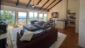 A spacious living room with a large brown sectional sofa, a white rug, and a kitchen area with white cabinets and a dining table.