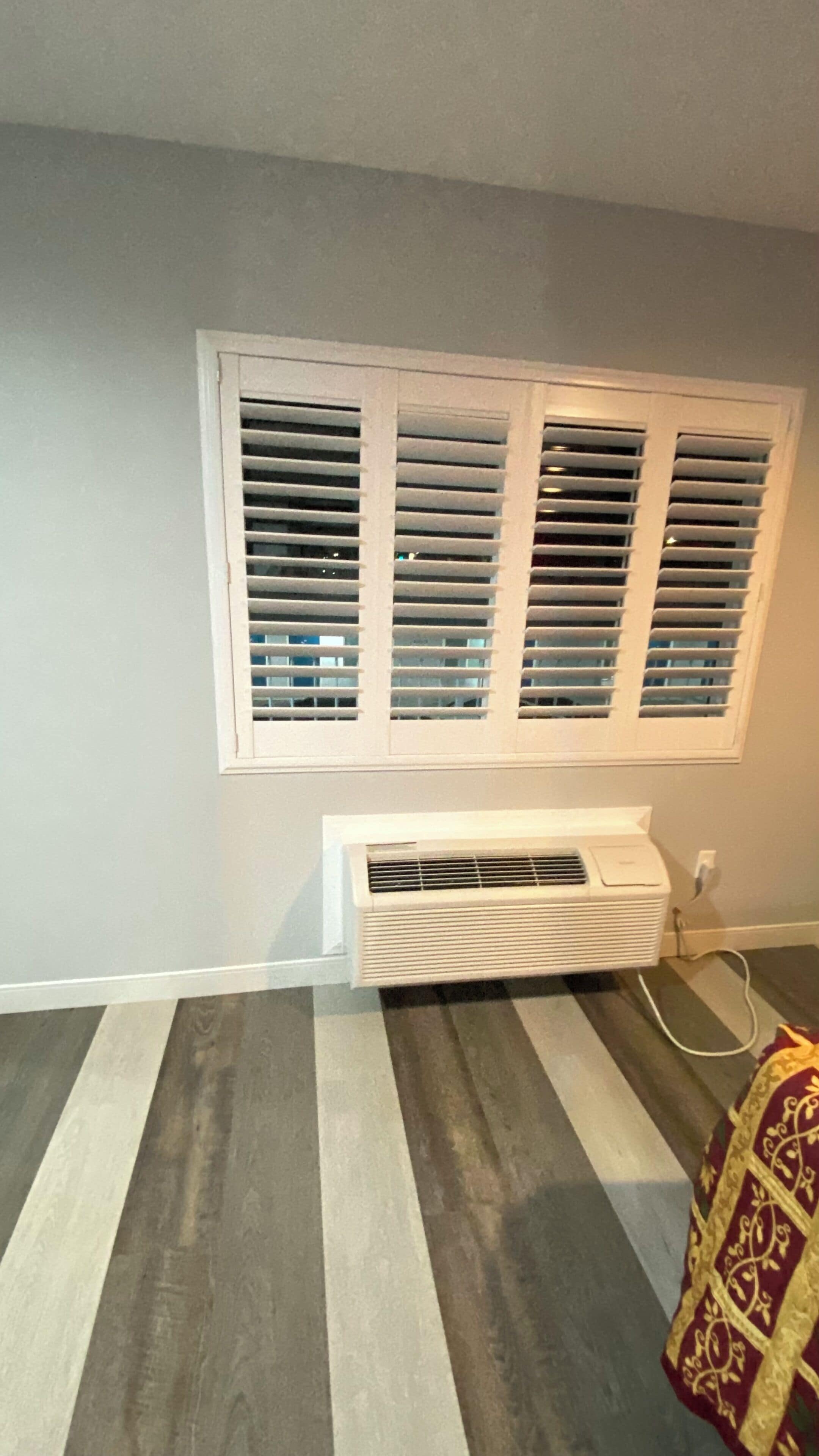 A room with a window with horizontal blinds, a white air conditioning unit below it, and a striped carpet on the floor.
