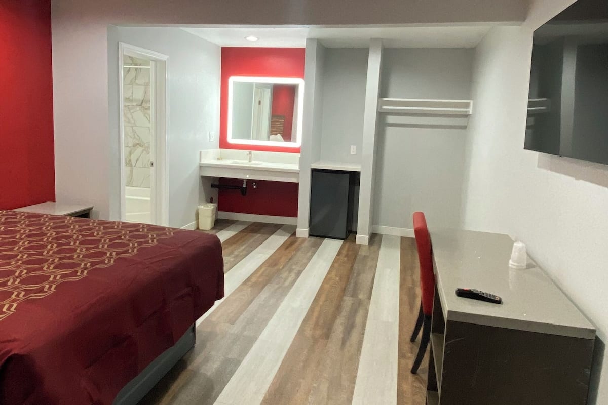 A hotel room with a bed, a desk, a chair, a TV, and a bathroom with a mirror and sink.