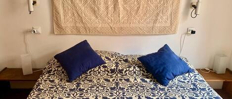 A bed with a patterned bedspread and two blue pillows, a beige curtain, and two wall-mounted light fixtures.