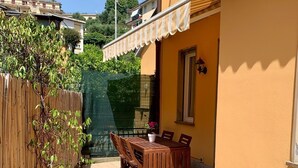 A patio with a wooden table and chairs, a small potted plant, and a building with a yellow exterior and a white awning. - Paola Cosy Apartment with A/C near best beaches (Monterosso al Mare)
