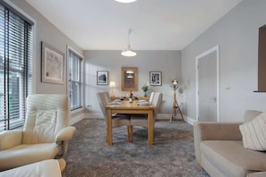 A dining area with a wooden table set for four, surrounded by chairs, a floor lamp, and framed pictures on the walls.