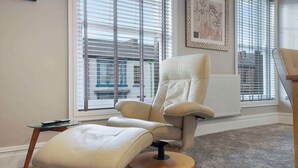 Una habitación con un gran ventanal, un sillón blanco, una mesita y un cuadro en la pared.