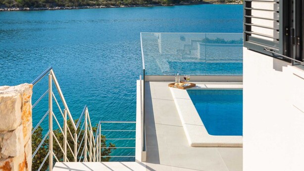 Ein Pool mit Glasgeländer und Blick auf ein Gewässer, ein Korbtablett mit Gläsern und Brot sowie eine Steinmauer.