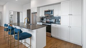 A modern kitchen with white cabinetry, stainless steel appliances, and a bar area with blue bar stools. - Niagara Falls Penthouse | Great Views | Gym | Luxe (Niagara Falls)