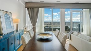 A dining area with a long table, chairs, a sofa, and a view of the sky through large windows. - Niagara Falls Penthouse | Great Views | Gym | Luxe (Niagara Falls)