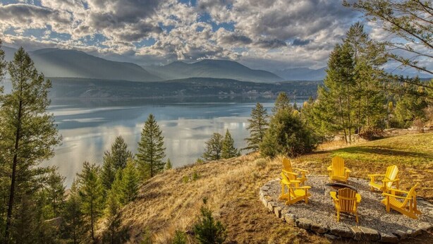 Une vue sur le lac, avec une rangée de chaises Adirondack jaunes faisant face à un lac paisible, le tout entouré de pins et avec des montagnes en arrière-plan.