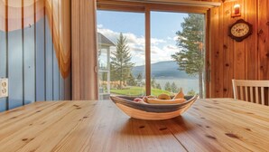 Une table à manger en bois, avec un saladier rempli de fruits au centre, une chaise en bois à droite, et une vue sur un lac et des montagnes, visible à travers une grande fenêtre.
