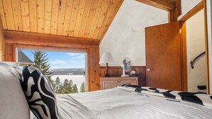 Une chambre à coucher avec un lit, une table de chevet, une lampe et un ventilateur. Il y a une fenêtre donnant sur des arbres et un plan d’eau.
