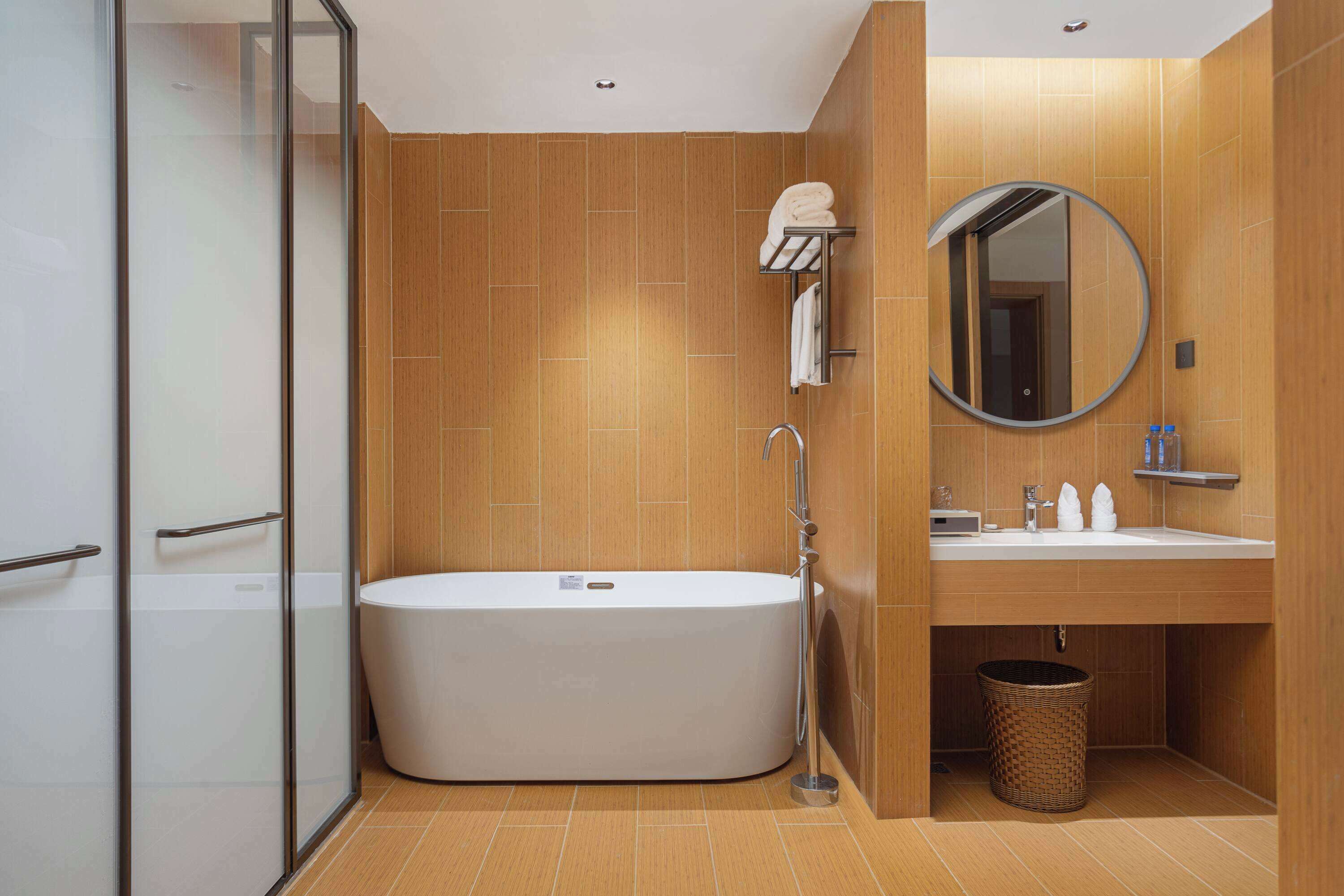 A modern bathroom with a freestanding bathtub, a glass shower enclosure, a wooden vanity with a sink and mirror, and a wicker waste basket.