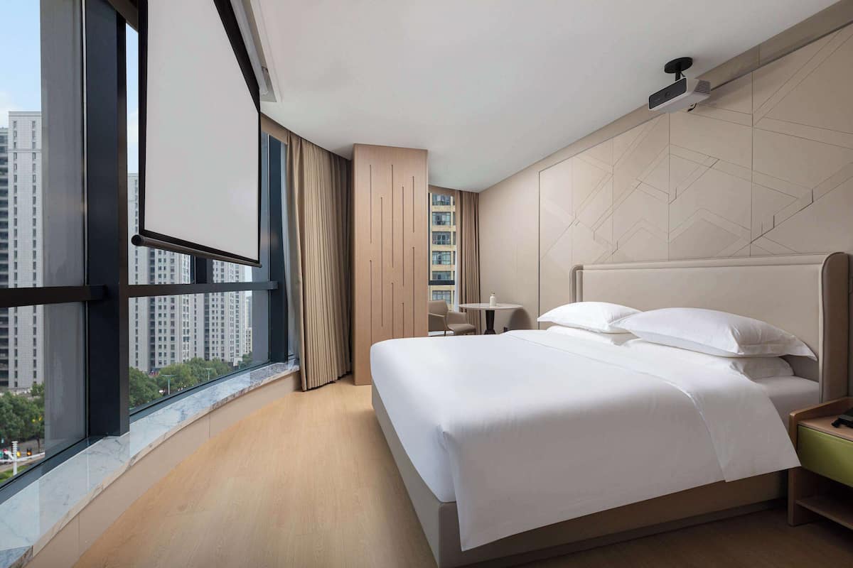 A hotel room with a large bed, a desk, a chair, and a view of tall buildings.